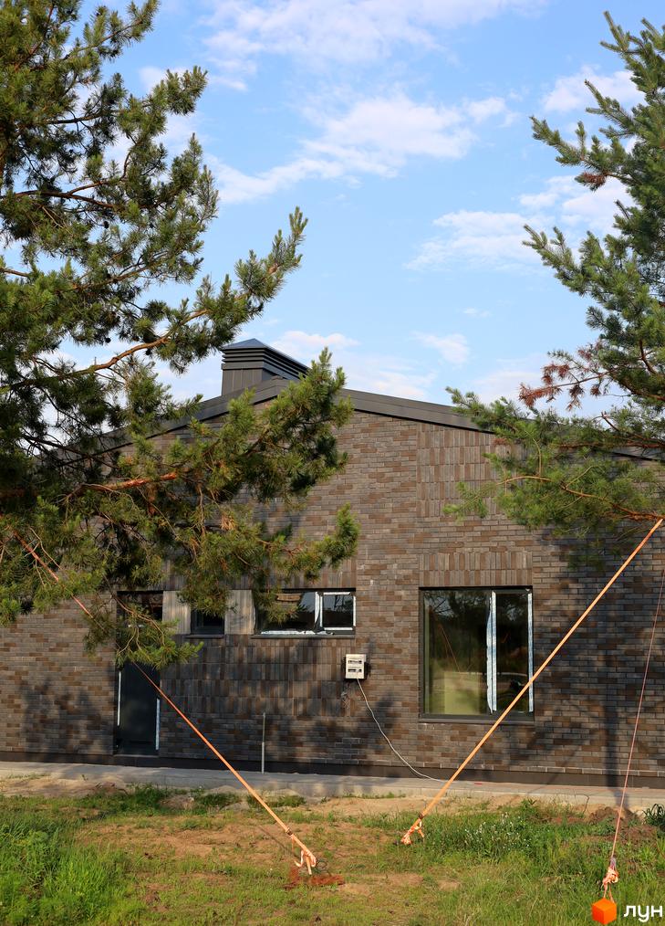 Котеджне містечко КМ NAVY Village з фасадом з темної цегли та великими вікнами. Навколо будинку зелена територія з деревами та травою.