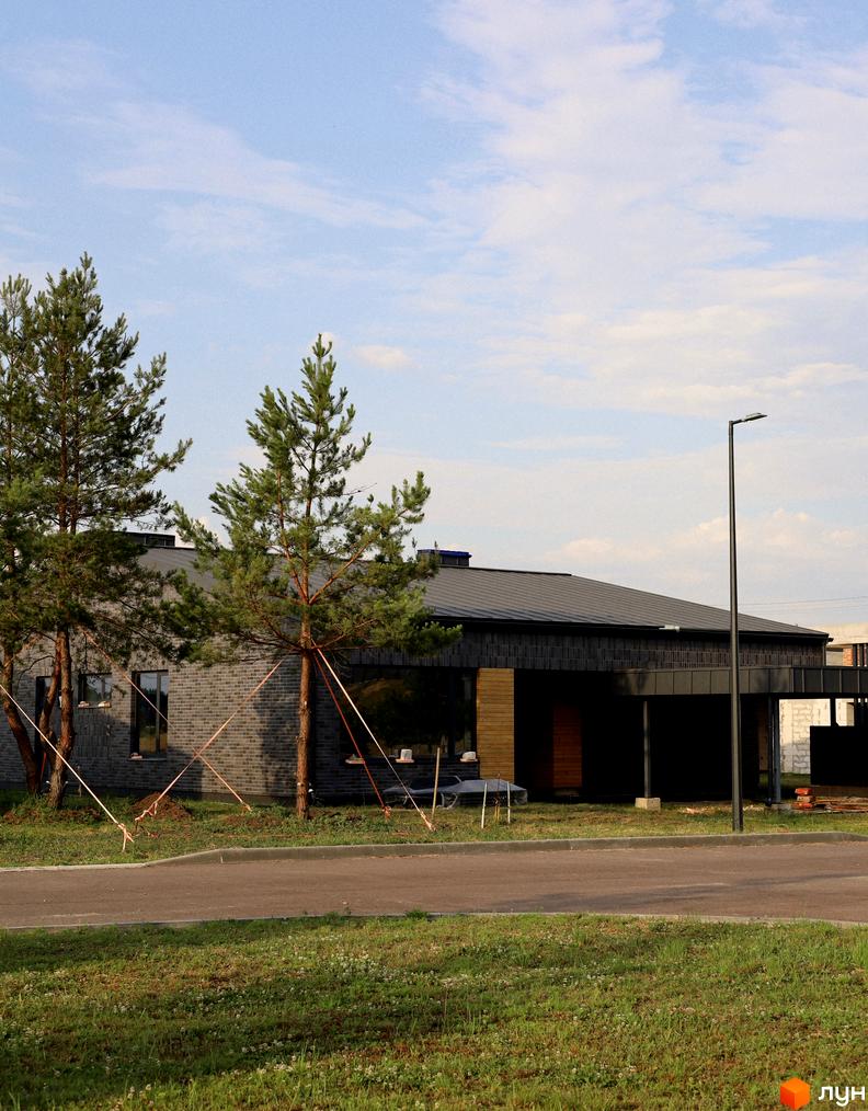 Котеджне містечко КМ NAVY Village з цегляним фасадом та великими вікнами, оточене соснами. Навколо будинку облаштована прибудинкова територія з дорогою та зеленою галявиною.