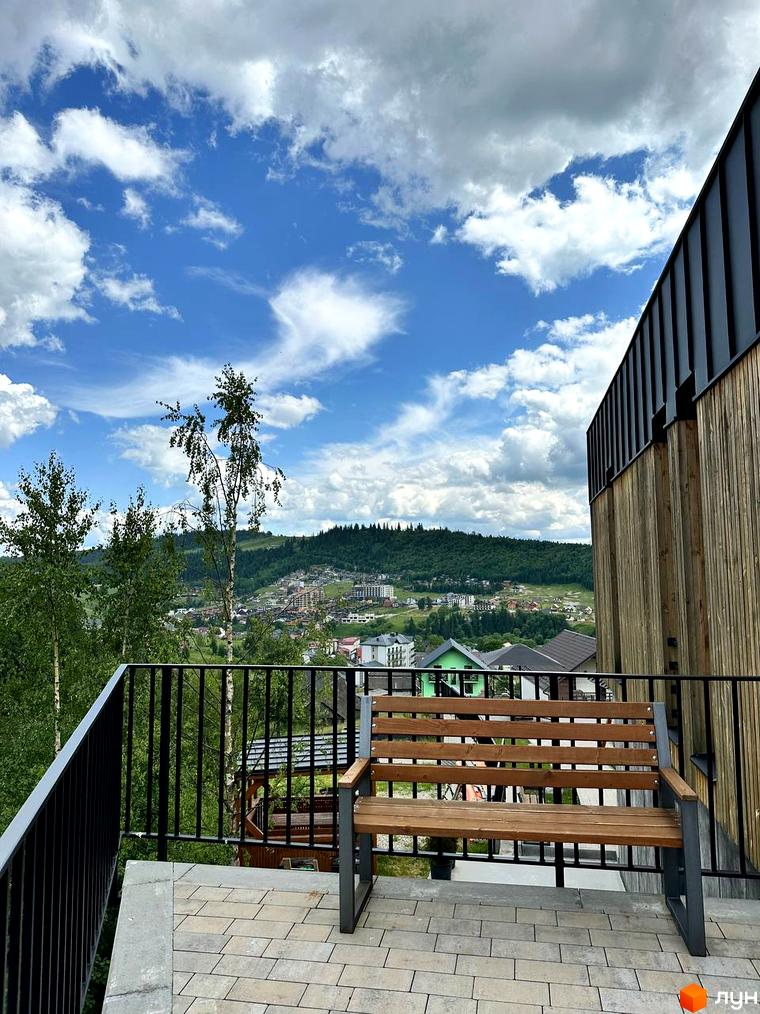 Апарт-готель Panorama Chalet з дерев'яною обшивкою та видом на зелені схили гір. На терасі встановлено лаконічну дерев'яну лавку.