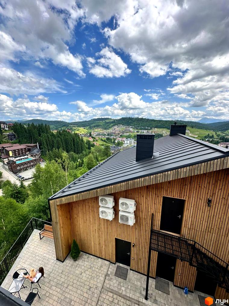 Апарт-готель Panorama Chalet з дерев'яним фасадом та чорним дахом, розташований на схилі гори з видом на зелені пагорби та хмарне небо. На території комплексу видно басейн та зелені насадження.