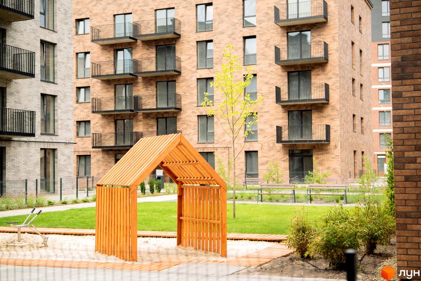 Житловий комплекс WAWEL з цегляним фасадом та балконами, поруч дитячий майданчик з дерев'яною альтанкою та зеленою зоною.