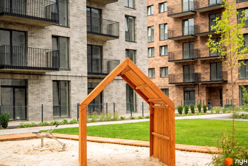 Житловий комплекс WAWEL з цегляними фасадами та балконами. На прибудинковій території пісочниця з дерев'яною аркою та зелений газон.