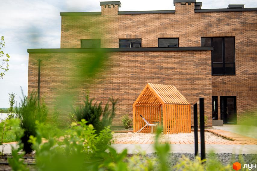 Будинок з цегляним фасадом та дерев'яною альтанкою на піщаній зоні. Територія новобудови ЖК WAWEL з елементами ландшафтного дизайну.