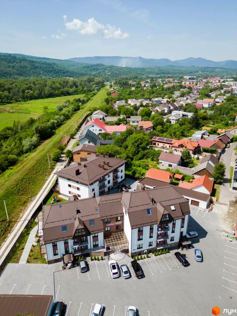 Житловий комплекс Comfort House з коричневим дахом та білими фасадами, розташований біля парковки з автомобілями та зеленої зони.