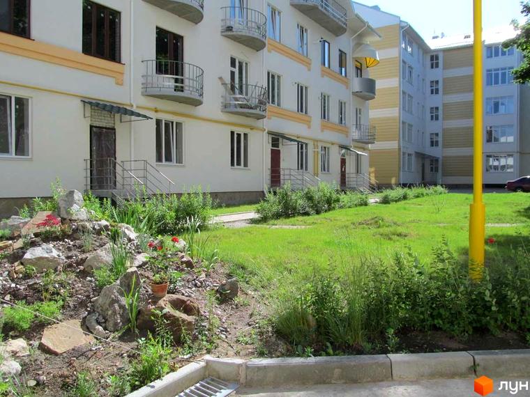 Житловий комплекс Околиця: біла фасадна частина будинку з балконами та вікнами. Прибудинкова територія з газоном та кам'яним ландшафтом.