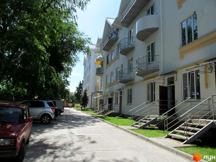 Зовнішній вигляд багатоквартирного будинку ЖК Околиця з балконами та сходами. Прибудинкова територія з автомобілями та деревами.