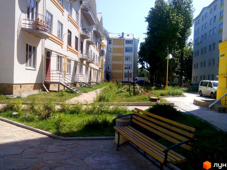 Зовнішній вигляд будинків житлового комплексу Околиця з прибудинковою територією. На зображенні видно фасади будівель, балкони та зелену зону з лавкою.