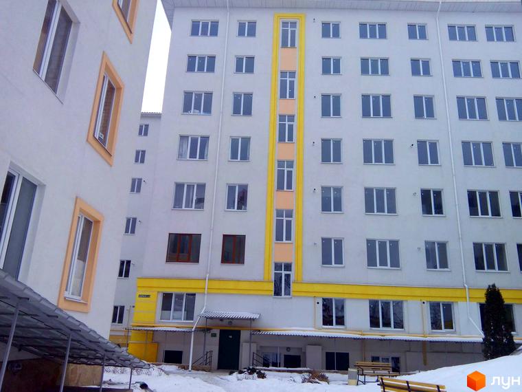 Зовнішній вигляд новобудови ЖК Околиця з численними вікнами та жовтими декоративними елементами. На прибудинковій території видно засніжену землю та лавки.