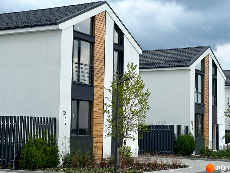 Котеджне містечко Gardd House Eco Village. Фасад будинку з білою штукатуркою, дерев'яними вставками та темними вікнами. Поруч паркан та зелені насадження.