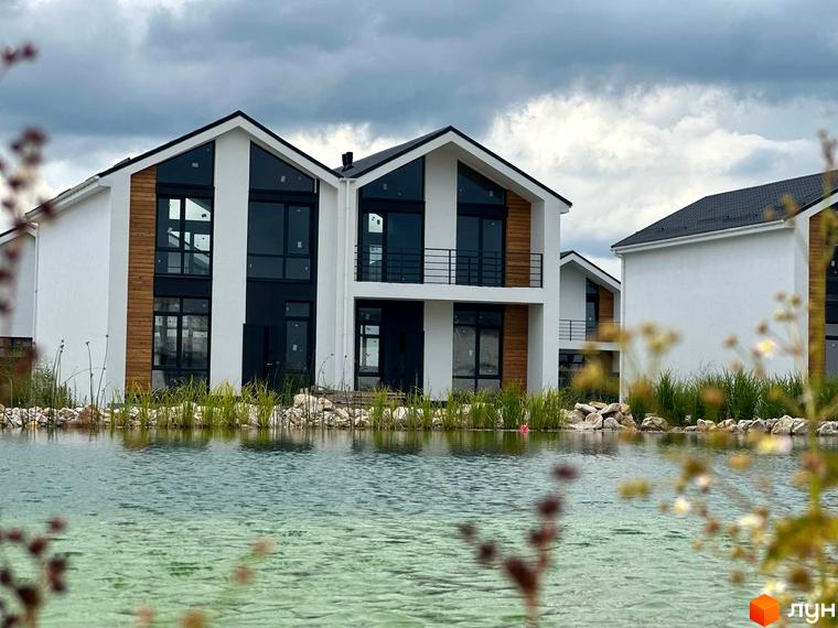 Котеджне містечко Gardd House Eco Village з білими фасадами, дерев'яними вставками та великими вікнами, розташоване біля водойми з зеленою травою та камінням.