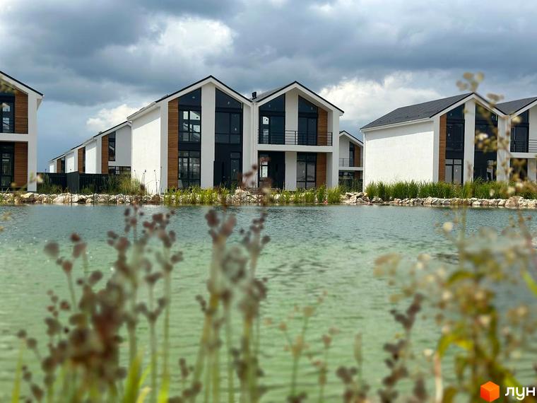 Котеджне містечко Gardd House Eco Village з білими фасадами, дерев'яними вставками та великими вікнами, розташоване біля водойми з зеленню.