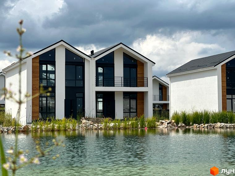 Котеджне містечко KМ Gardd House Eco Village з білими фасадами, дерев'яними вставками та великими вікнами, розташоване біля водойми з зеленою травою.