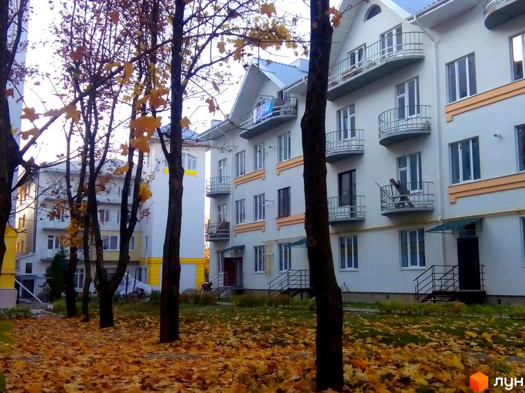 Зовнішній вигляд новобудови ЖК Околиця з балконами та вікнами на тлі осінніх дерев та опалого листя. Прибудинкова територія з газоном та деревами.