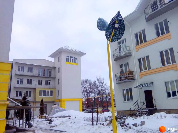 Зовнішній вигляд новобудови ЖК Околиця з балконами та вікнами. На прибудинковій території видно ліхтар у вигляді листя та засніжену землю.
