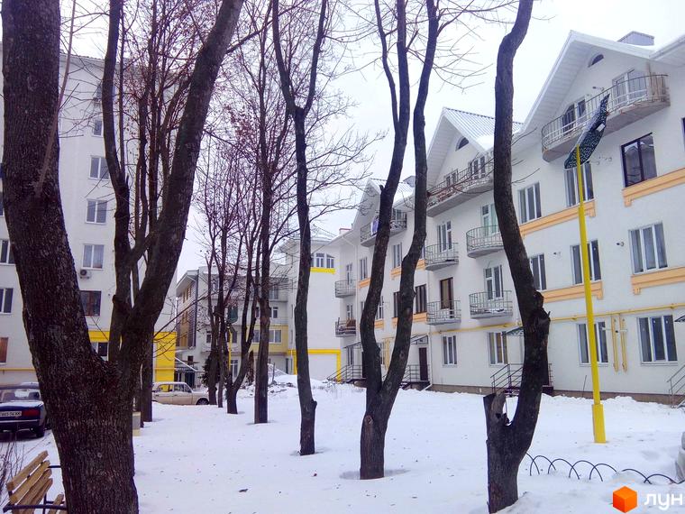 Житловий комплекс Околиця. Біла будівля з жовтими елементами та балконами. Навколо дерева та засніжена територія.