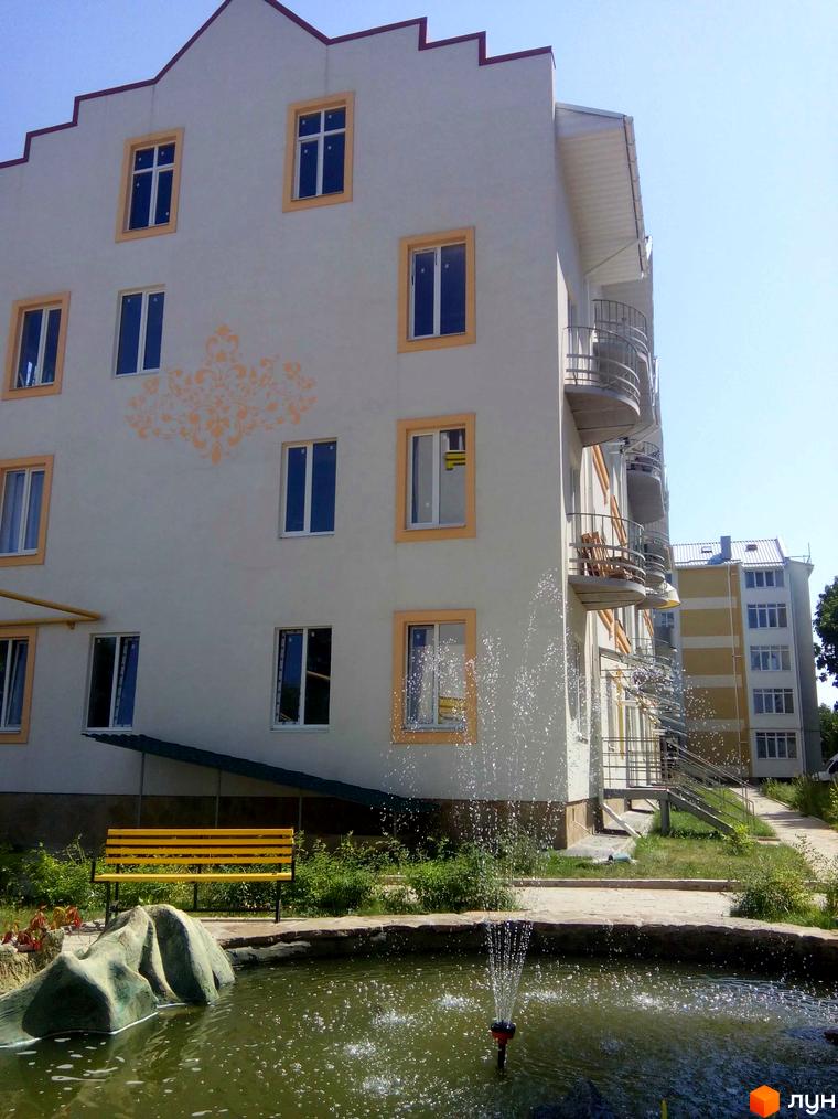 Житловий комплекс Околиця. Фасад будинку з вікнами та балконами, поруч фонтан та лавка.