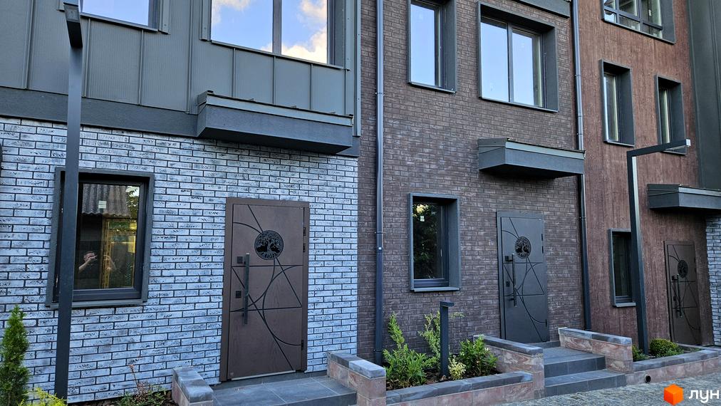 Фасад таунхаусів New Smart 17 з цегляною обробкою та дверима з візерунком дерева. Прибудинкова територія з рослинами та сходами.