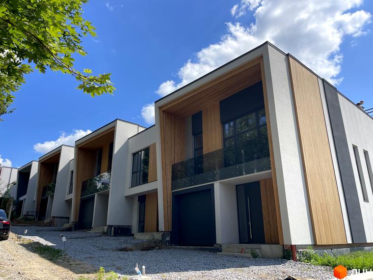 Таунхауси FIRST – сучасні будинки з дерев'яними елементами та скляними балконами на тлі блакитного неба. Навколо новобудов – гравійне покриття та зелені насадження.
