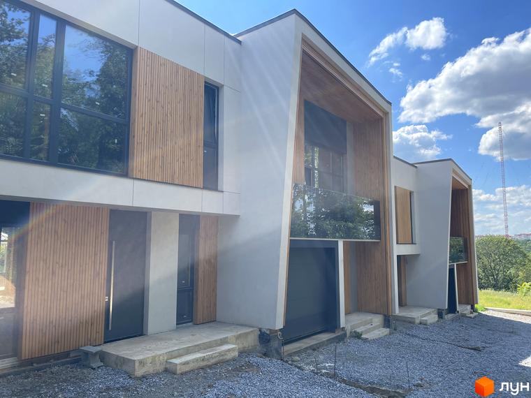 Таунхауси FIRST: фасади з дерев'яними елементами та великими вікнами, перед будинками гравій та бетонні сходи. Навколо зелені насадження та ясне небо з хмарами.