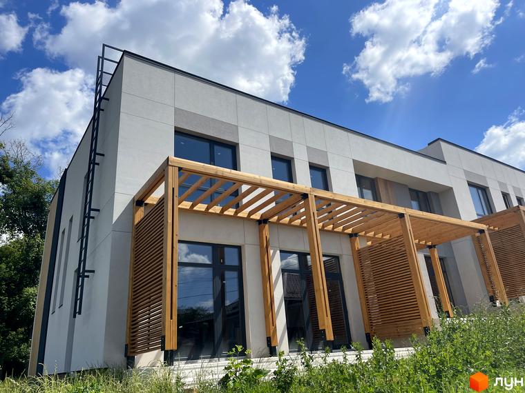 Таунхауси FIRST з дерев'яною перголою та ламелями на фасаді. Зовнішній вигляд новобудови на фоні блакитного неба з хмарами.
