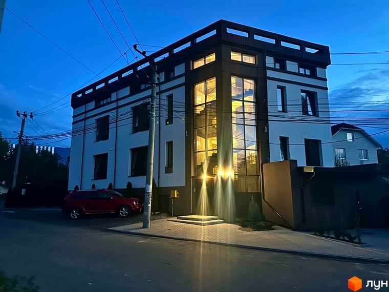 Житловий комплекс Olympia house з панорамними вікнами та прибудинковою територією ввечері. Червоний автомобіль припаркований біля будинку.