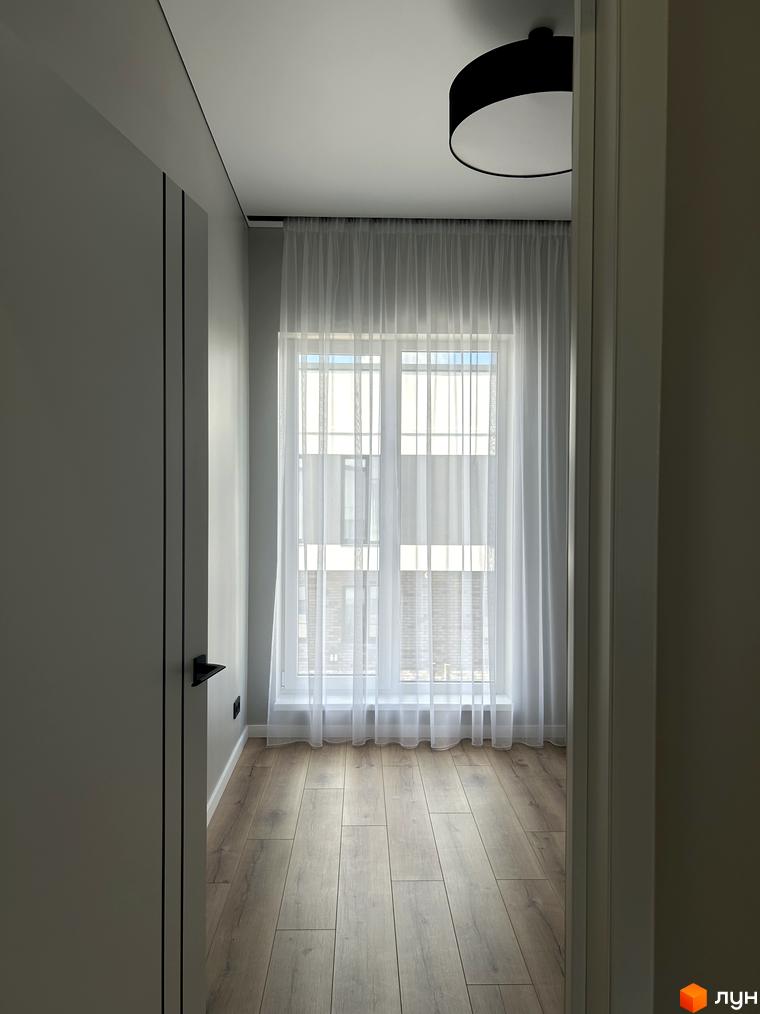 Світла кімната з великим вікном, затягнутим прозорим тюлем, та дерев'яною підлогою. На стелі розташований чорний світильник.