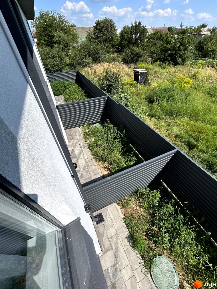 Фасад таунхауса Peremohy Homes з сірим профільним парканом та вимощеною доріжкою. Навколо зелена територія з травою та деревами.