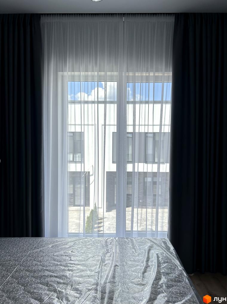 Вікно з білими напівпрозорими шторами, за яким видно сучасну двоповерхову будівлю з великою кількістю вікон та сірим дахом.