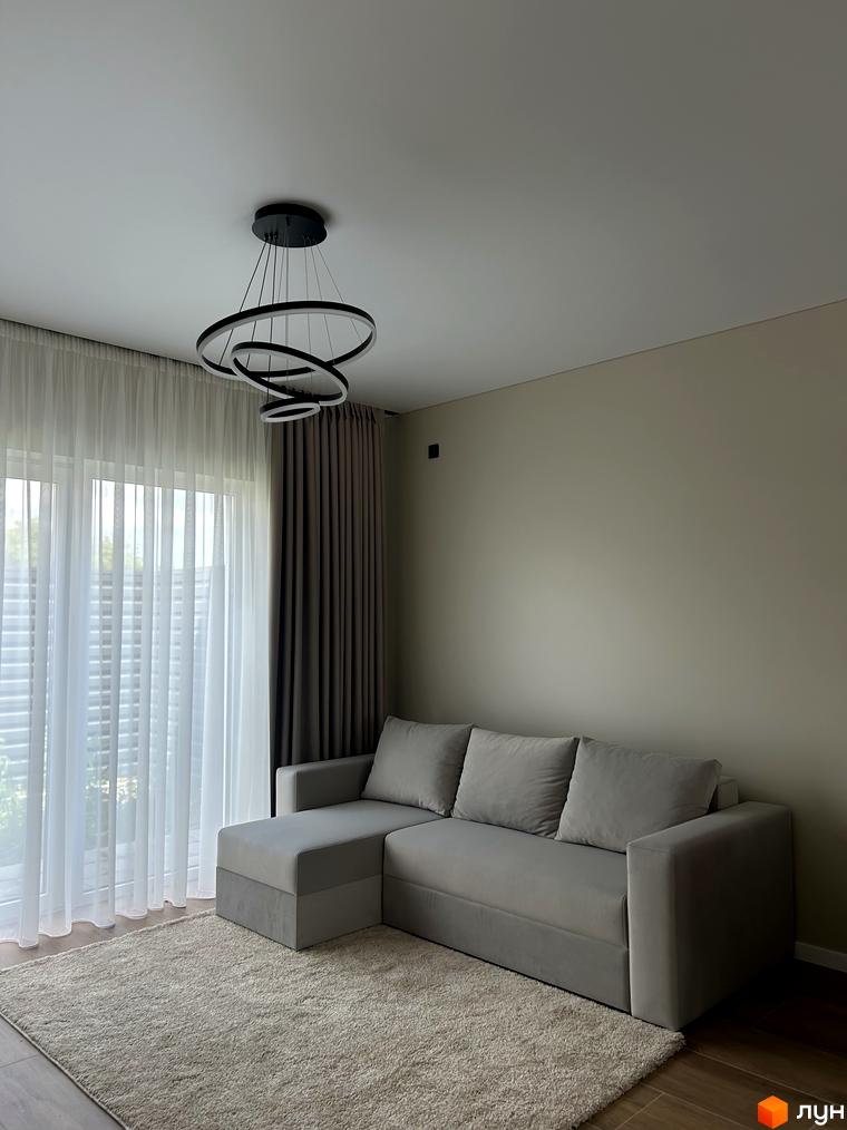 Вітальня в Peremohy Homes з диваном, килимом та багатоярусною люстрою. Великі вікна з білими напівпрозорими шторами забезпечують багато природного світла.