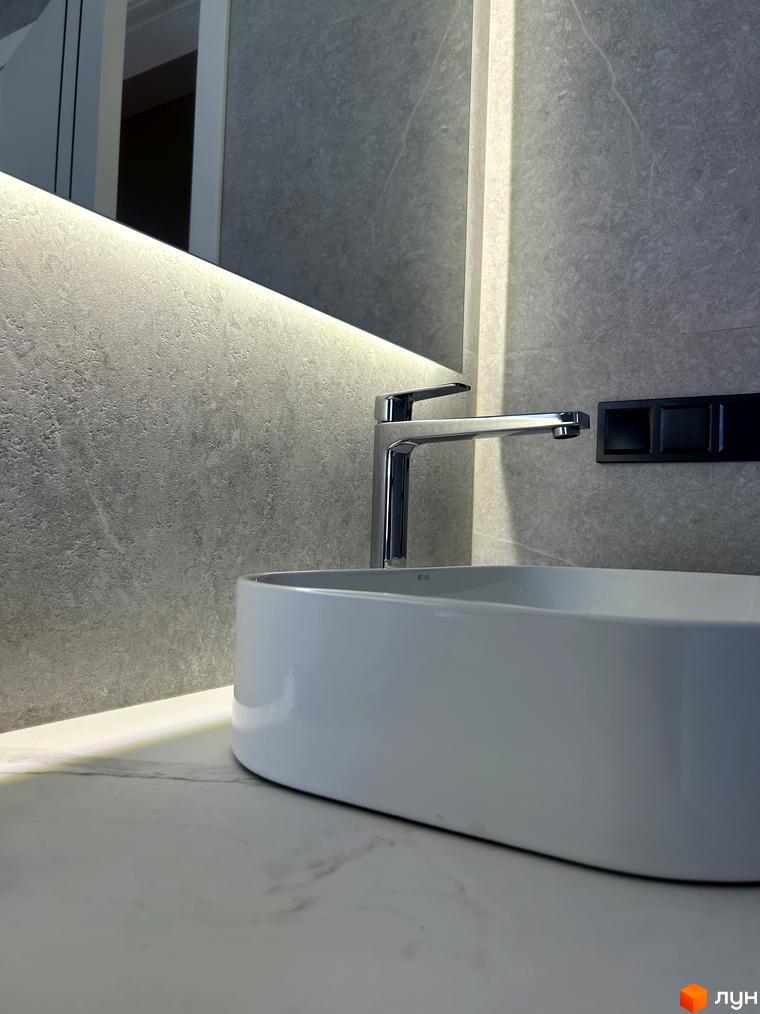 Сучасний дизайн ванної кімнати з білою раковиною та хромованим змішувачем на тлі сірої плитки з підсвічуванням.