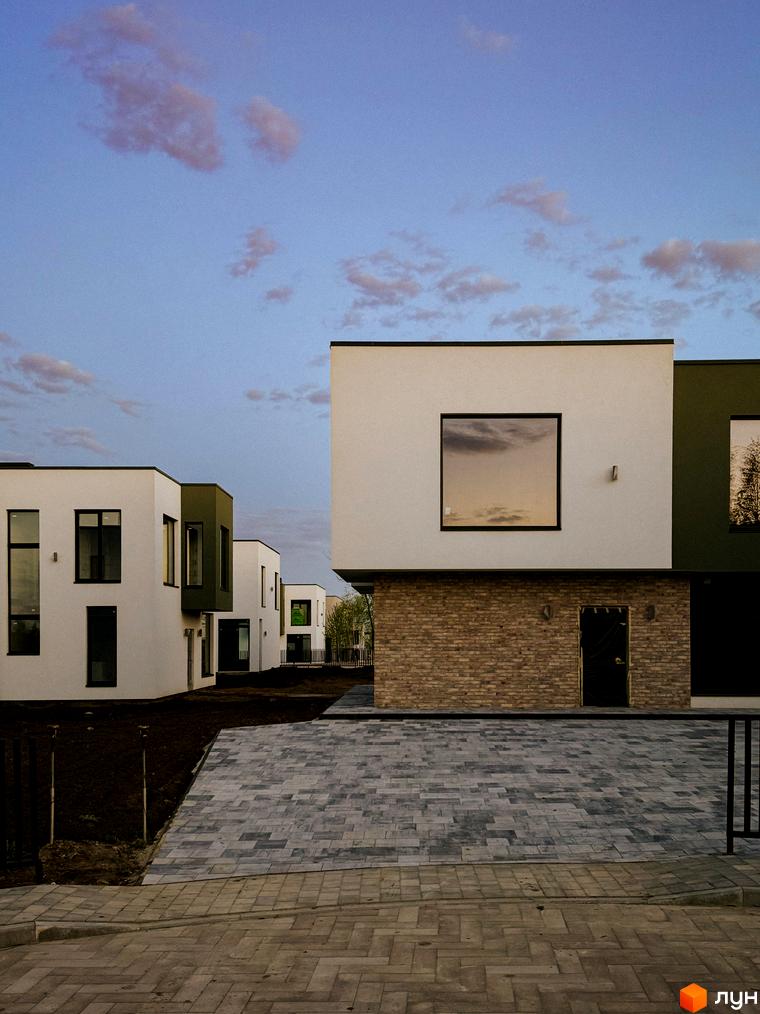 Котеджне містечко Barcelona: білі будинки з темними вікнами та зеленими вставками, вимощена сіра доріжка перед ними.