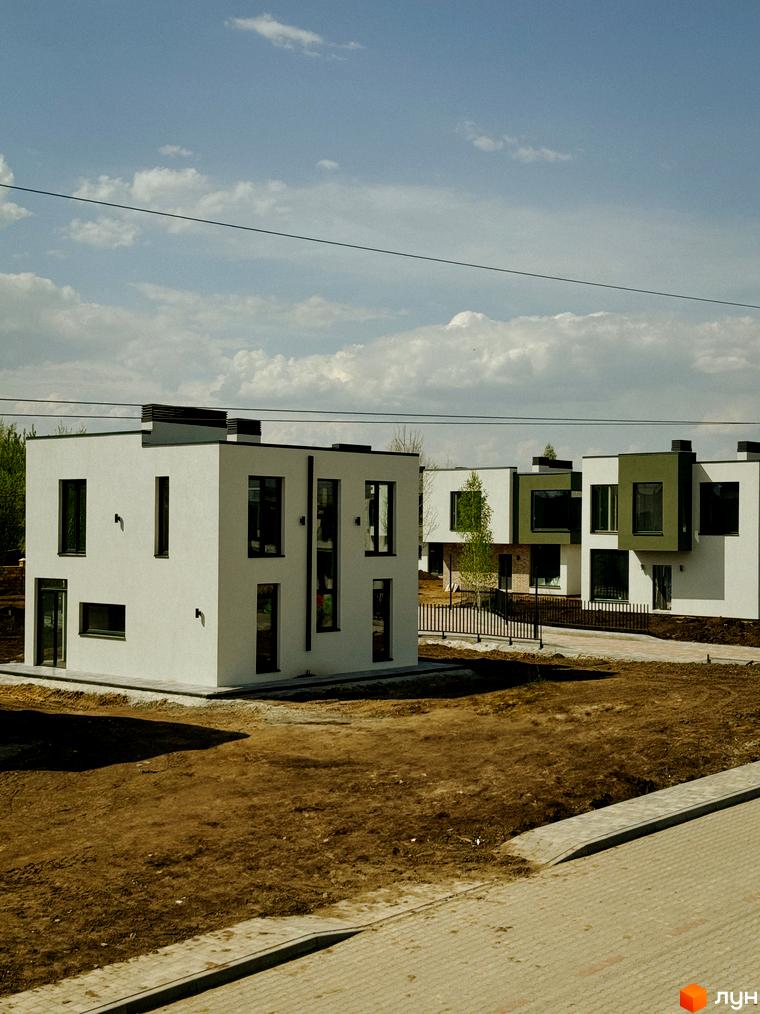 Котеджне містечко Barcelona: білі двоповерхові будинки з чорними вікнами та зеленою обробкою, на тлі блакитного неба з хмарами. Навколо будівельний майданчик з плиткою та ґрунтом.