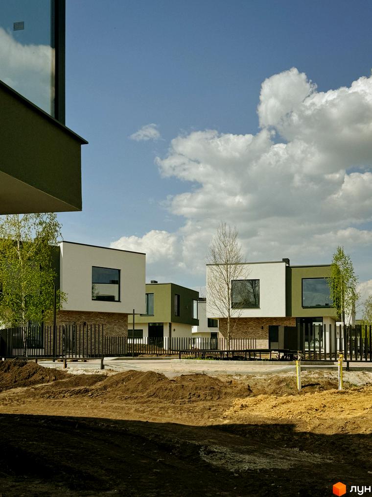 Котеджне містечко Barcelona: фасади будинків з білою та зеленою штукатуркою, великими вікнами. Територія з парканом, деревами та ділянкою з будматеріалами.