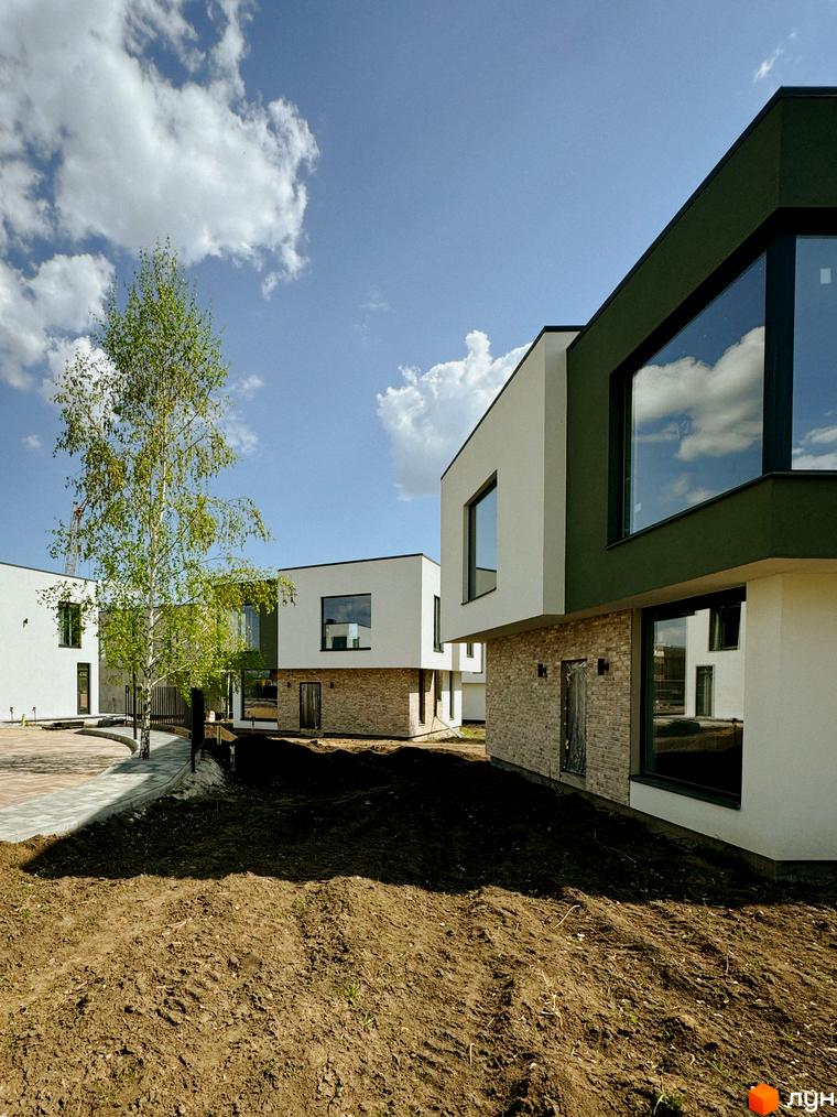 Котеджне містечко Barcelona: білі та зелені фасади будинків з великими вікнами, вимощена доріжка та зелене дерево на фоні блакитного неба з хмарами.