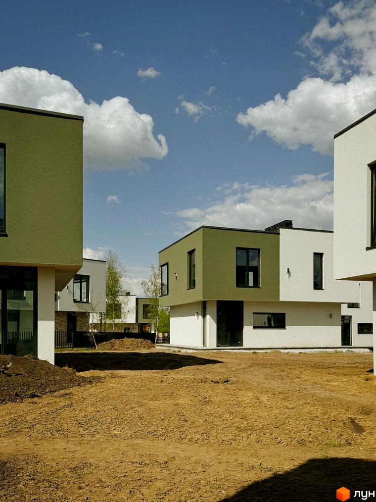 Котеджне містечко Barcelona: сучасні будинки з зеленими та білими фасадами на тлі блакитного неба з хмарами. Навколо новобудов - ділянка з ґрунтом та молодими деревами.