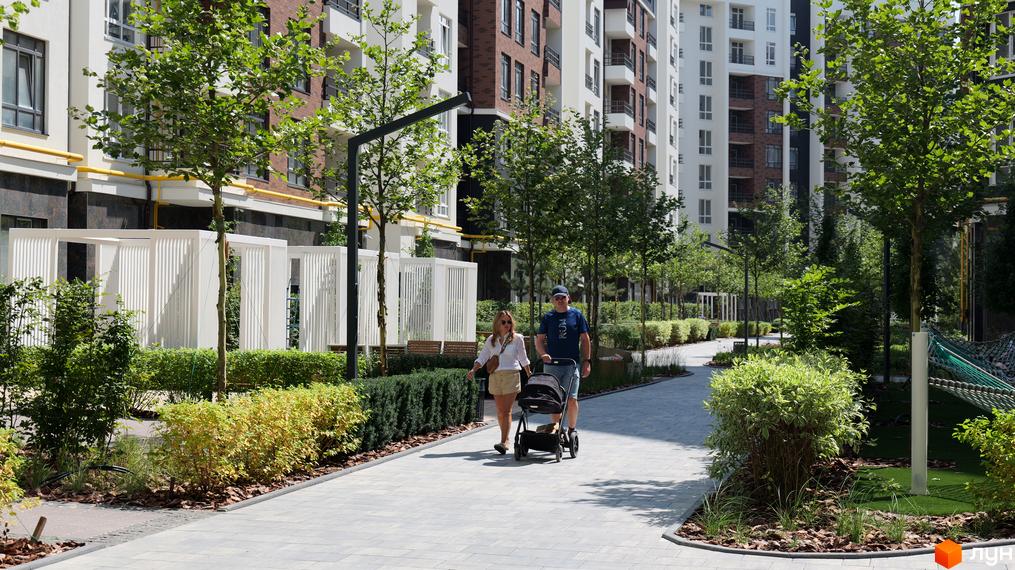 Житловий комплекс Синергія Сіті з новими будинками та зеленою прибудинковою територією, де гуляють люди з дитячим візочком.