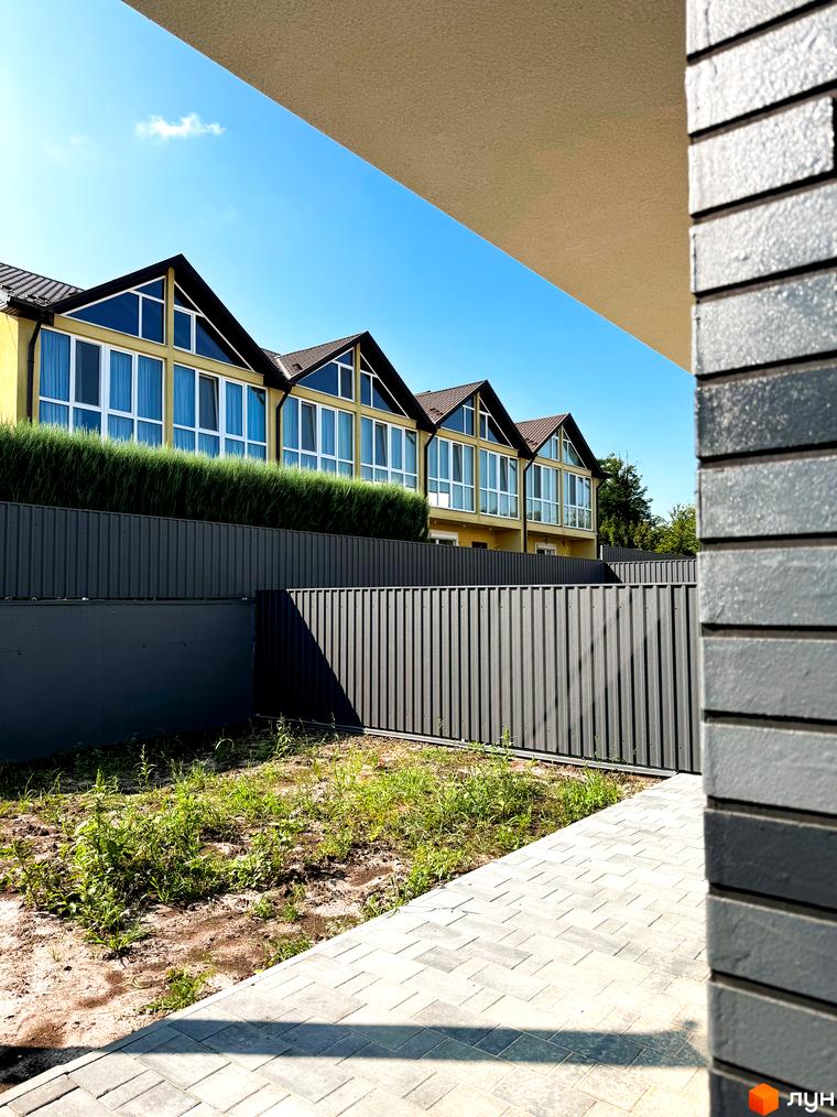 Зовнішній вигляд новобудови на вул. Троянівська, 21а, з великими вікнами та зеленим парканом. Прибудинкова територія з бруківкою та травою.