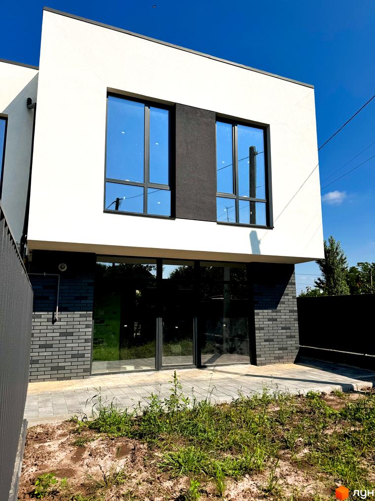 Сучасний двоповерховий будинок з великими вікнами та сірою цегляною обробкою першого поверху. Територія перед будинком вимощена плиткою, засаджена зеленню.