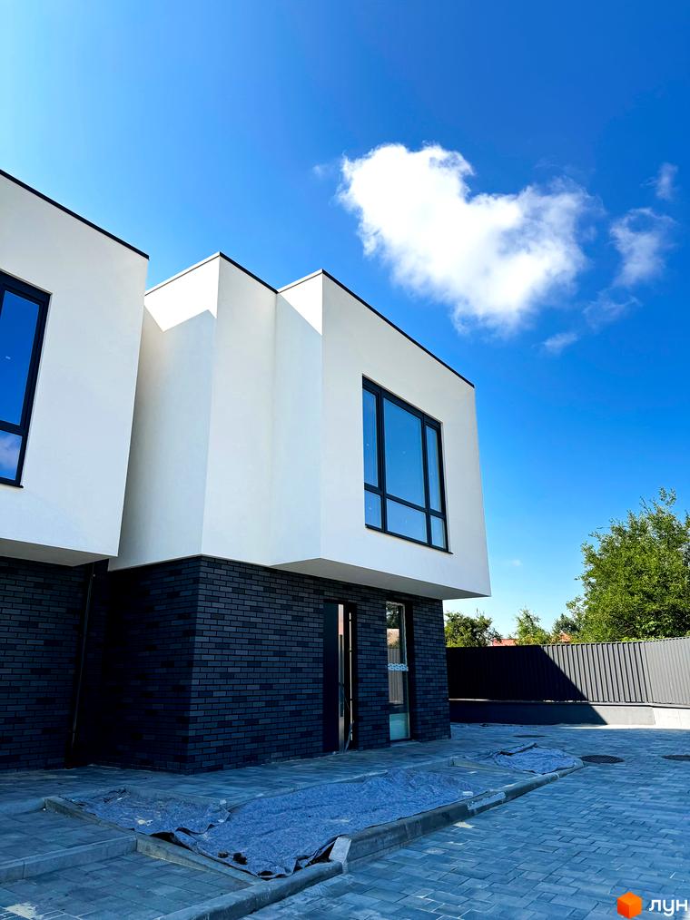 Фасад новобудови на вул. Троянівська, 21а, з білими стінами та темною цегляною основою. Прибудинкова територія вимощена плиткою.