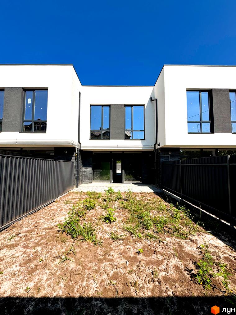 Фасад новобудови на вул. Троянівська, 21а, з великими вікнами та чорною огорожею. Попереду ділянка з травою та піском.