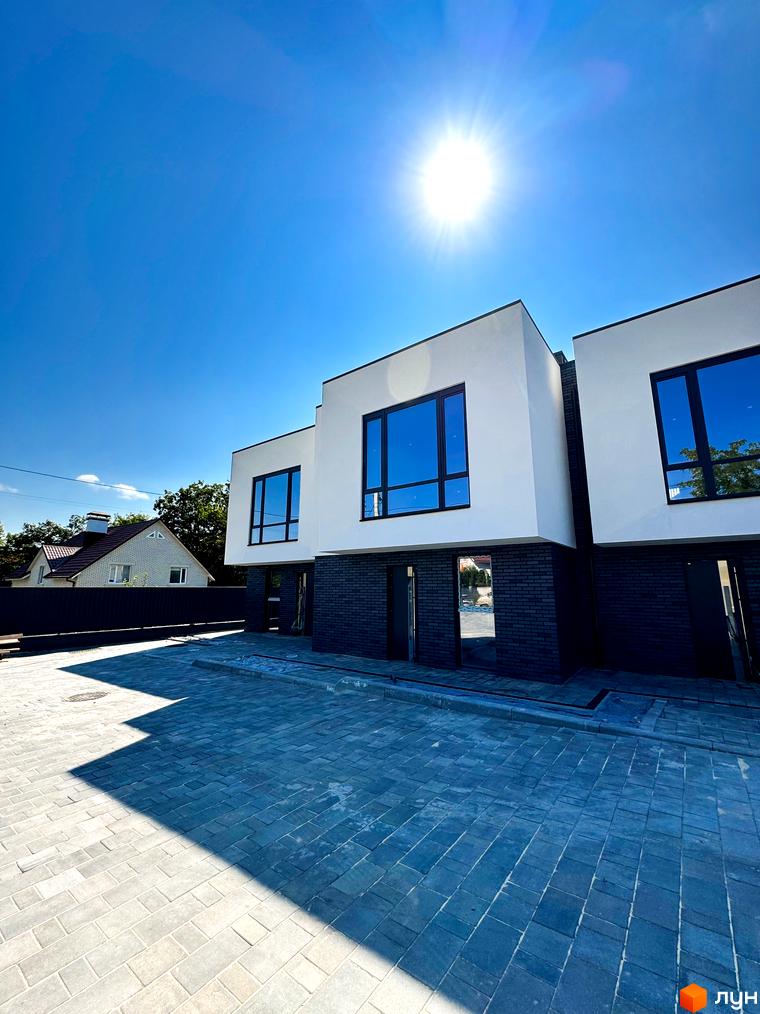 Будівельний проєкт на Троянівській, 21а: триповерхові будинки з білою штукатуркою та темною цеглою, великими вікнами, на фоні блакитного неба. Прибудинкова територія з бруківки.