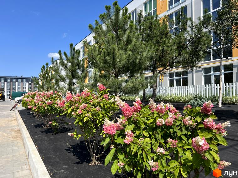 Житловий комплекс Sanville Park з квітучими кущами та деревами на фоні будівель під блакитним небом.