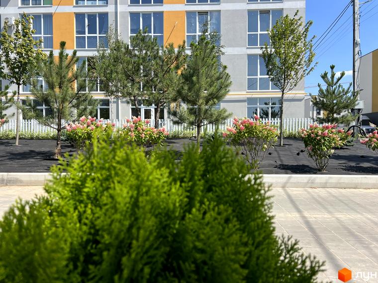 Фасад житлового комплексу Sanville Park з елементами оранжевого кольору та зеленою прибудинковою територією з квітучими кущами.