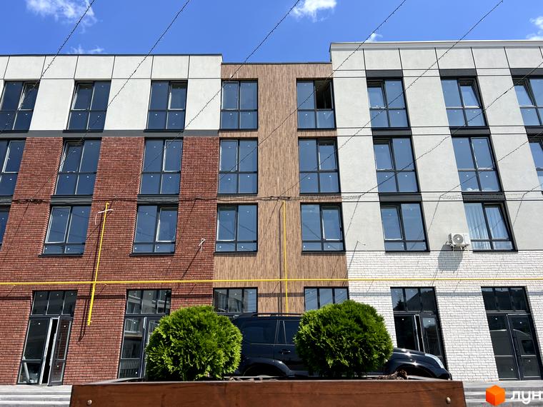 Фасад житлового комплексу Sanville Park з цегляними та штукатуреними елементами, великими вікнами та зеленими кущами перед будинком.