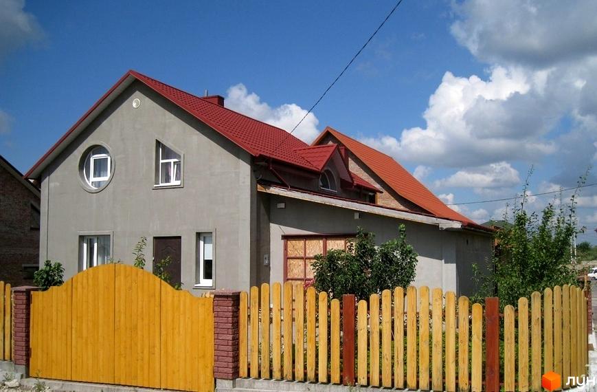 Зовнішній вигляд будинку з червоним дахом та сірими стінами, вікнами різної форми та дерев'яним парканом. Комплекс КМ Кленовий гай – це затишна новобудова.