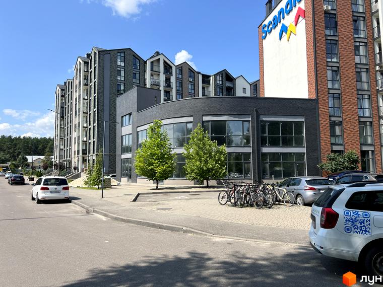 Зовнішній вигляд житлового комплексу Scandia з багаторівневими фасадами та панорамними вікнами. На прибудинковій території розташовані дерева та парковка з велосипедами.