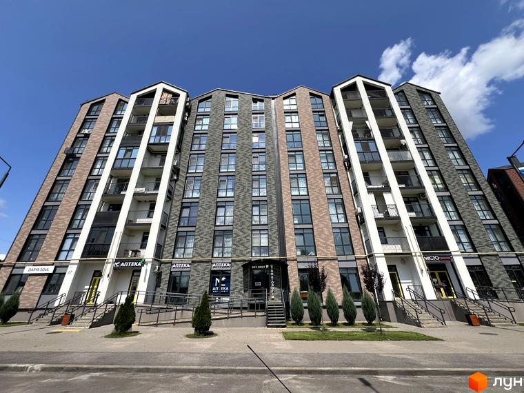 Фасад житлового комплексу Scandia з цегляним оздобленням та балконами на тлі блакитного неба. Перед будинком розташована доглянута прибудинкова територія з деревами та доріжками.