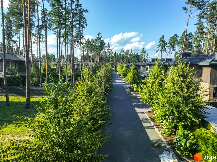 Котеджне містечко ZAHRAVA village 2: асфальтована дорога, що веде до будинків, оточена зеленими деревами та газонами. Навколо розташовані сучасні котеджі під сірими дахами.