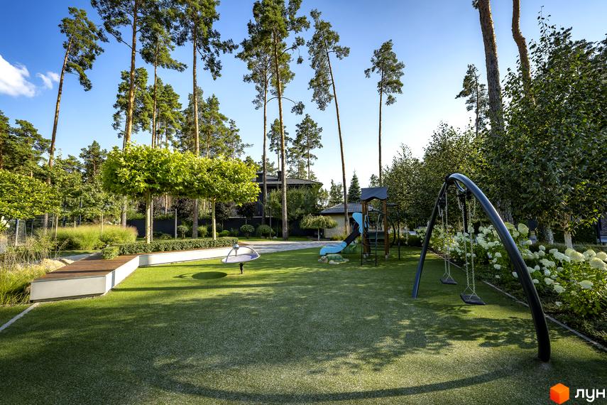 Котеджне містечко ZAHRAVA village 2. Дитячий майданчик з гойдалками, гіркою та каруселлю на фоні зеленої галявини та високих дерев.