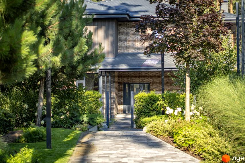 Котеджне містечко ZAHRAVA village 2. Фасад будинку з цегли та дерева, доріжка з бруківки веде до входу, навколо зелені насадження.